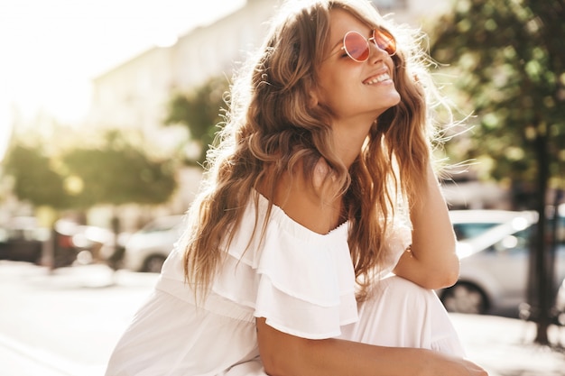 Porträt des schönen niedlichen blonden Teenagermodells ohne Make-up in den weißen Kleidungsstücken des Sommer-Hipsters, die auf dem Straßenhintergrund sitzen