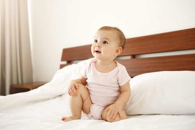 Porträt des schönen niedlichen netten Neugeborenen im rosa Hemd, das auf Bett zu Hause sitzt.