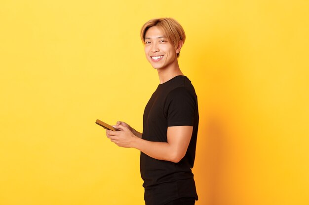 Porträt des schönen stilvollen asiatischen Kerls im schwarzen Outfit, mit Handy und drehendem Kopf mit zufriedenem Lächeln, gelbe Wand