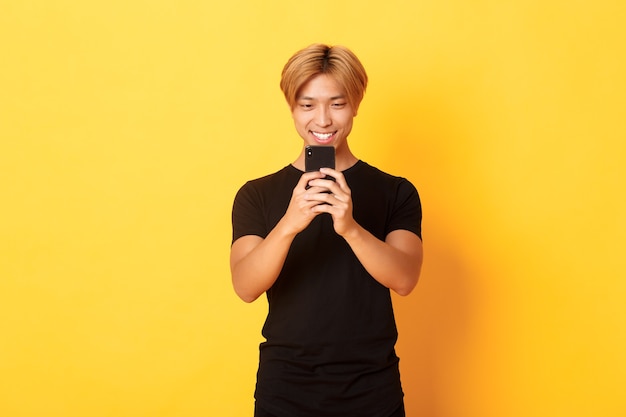 Porträt des schönen stilvollen asiatischen kerls mit blondem haar, unter verwendung des mobiltelefons und des lächelns, nachrichten in der smartphone-app, gelbe wand