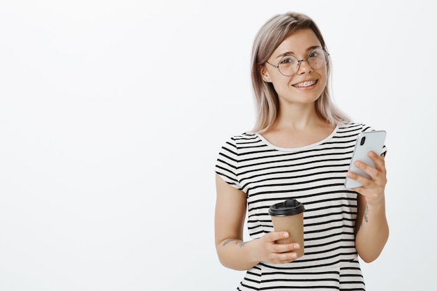 Porträt des selbstbewussten blonden Mädchens, das im Studio mit ihrem Telefon und Kaffee aufwirft