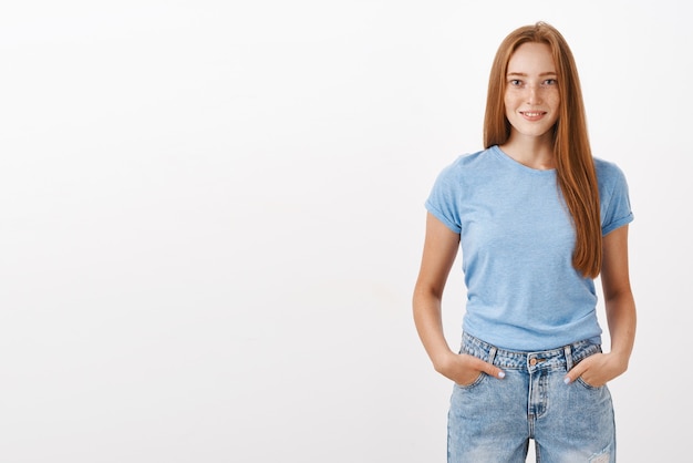 Porträt des selbstbewussten gut aussehenden Ingwermädchens mit niedlichen Sommersprossen im blauen T-Shirt und in den Jeans, die Hände in den Taschen halten und mit selbstbewusstem Ausdruck lächeln