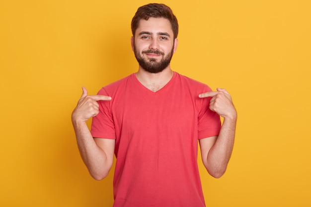 Porträt des selbstbewussten gut aussehenden kaukasischen Mannes, der rotes lässiges T-Shirt trägt, über gelb steht und auf sein T-Shirt zeigt