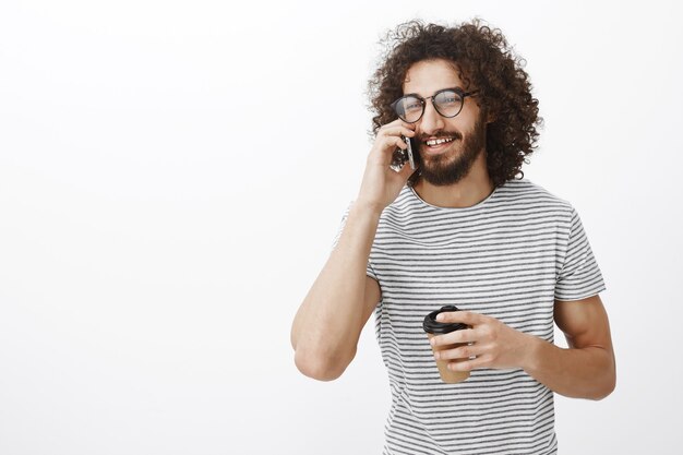 Porträt des sorglosen gutaussehenden bärtigen Mannes in der Brille, der auf dem Smartphone spricht und mit freudigem Lächeln beiseite schaut und eine Tasse Kaffee oder Tee hält
