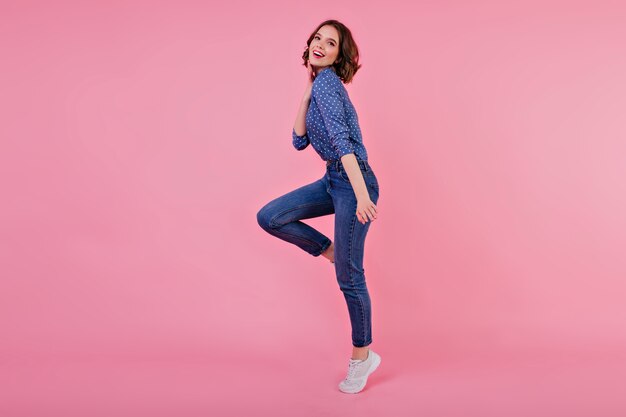 Porträt des sportlichen Mädchens in voller Länge mit gewelltem Haar. Innenaufnahme der springenden jungen Frau in den Jeans und im blauen Hemd.