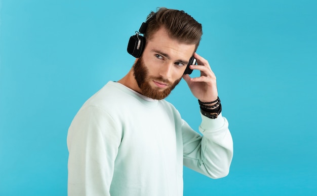 Porträt des stilvollen attraktiven jungen bärtigen Mannes, der Musik auf drahtlosen Kopfhörern des modernen Stils selbstbewusster Stimmung lokalisiert auf blauem Hintergrund hört