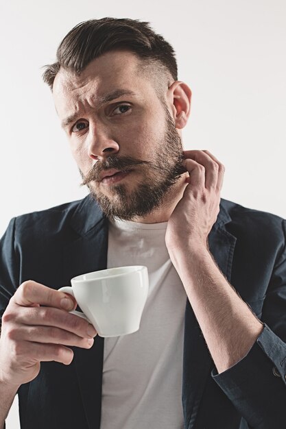 Porträt des stilvollen hübschen jungen Mannes, der am Studio steht. Mann, der Jacke trägt und Tasse Kaffee hält