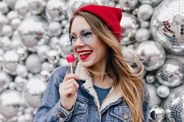 Porträt des stilvollen Mädchens in der Jeansjacke, die Süßigkeiten isst und wegschaut. Wunderbare europäische Dame im roten Hut und in den runden Gläsern, die mit Lutscher auf glänzender Wand aufwerfen.