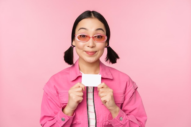 Porträt des stilvollen modernen asiatischen mädchens zeigt ermäßigte kreditkarte und sieht erfreut aus, kontaktloses konzept des einkaufens zu bezahlen, das über rosa hintergrundkopienraum steht