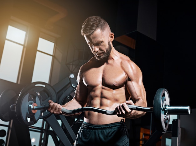 Porträt des super fit muskulösen jungen Mannes, der im Fitnessstudio mit Langhantel trainiert