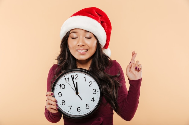 Porträt des Träumens des Mädchens in rotem Hut Santa Clauss, der die Uhr zeigt fast 12 macht Wunsch mit den Fingern hält, kreuzte über Pfirsichhintergrund