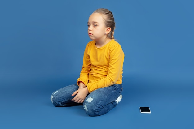 Porträt des traurigen kleinen Mädchens, das lokal auf blauem Hintergrund sitzt.