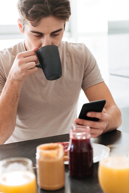 Porträt des trinkenden Tees des jungen fokussierten Mannes und der Anwendung des Smartphone