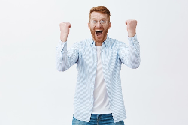 Porträt des triumphierenden glücklichen kaukasischen Mannes mit Ingwerhaar und Borsten in der Brille, die vom Erfolg und von den positiven Gefühlen schreien, die Fäuste erheben, um Sieg zu feiern