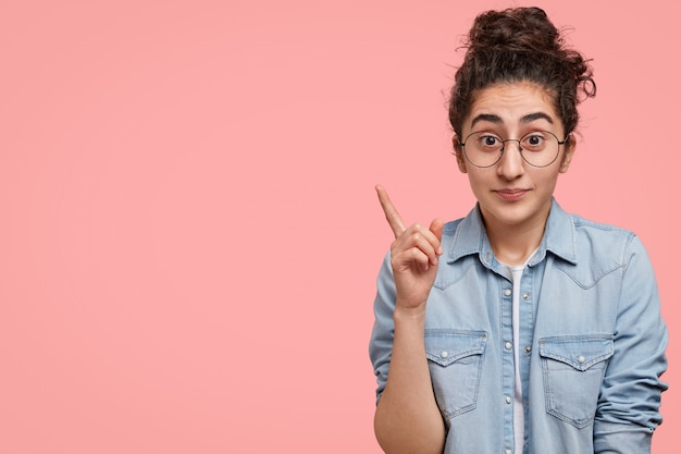 Porträt des überraschten brünetten kaukasischen weiblichen aussehens mit verwirrtem ausdruck, hebt den vorderfinger an der linken oberen ecke gegen eine rosa wand mit kopierraum