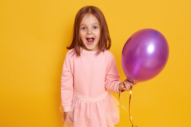 Porträt des überraschten kleinen Mädchens mit dunklem glattem Haar, das über gelben Studio schönen Kleidern steht und lila Ballon in den Händen hält