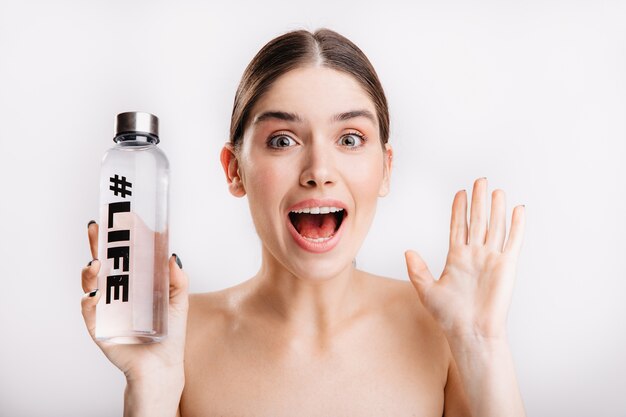 Porträt des überraschten Mädchens mit sauberer gesunder Haut. Dame posiert überrascht und hält eine Flasche Wasser mit Inschriftenleben darauf.