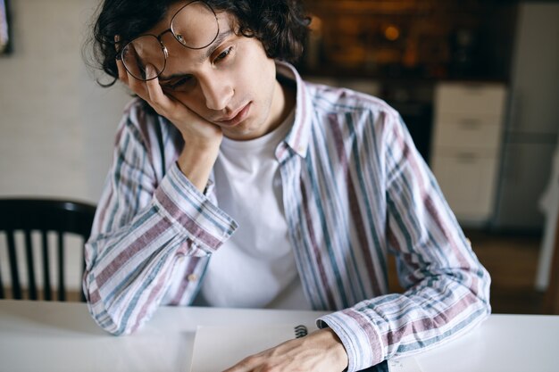 Porträt des verschlafenen männlichen Studenten in der Freizeitkleidung, die am weißen Schreibtisch sitzt, Hand auf seinem Gesicht hält, gelangweilten Blick hat, müde ist, Hausaufgaben zu machen, braucht etwas Schlaf.