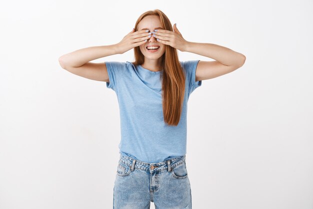 Porträt des verspielten und glücklichen jungen Mädchens der Rothaarigen im blauen lässigen T-Shirt, das Augen mit Handflächen bedeckt und beim Verstecken lächelt