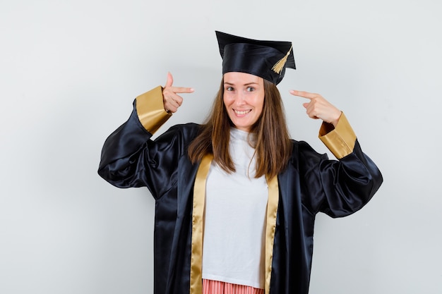 Porträt des weiblichen Absolventen, der auf ihren Kopf in der akademischen Kleidung zeigt und fröhliche Vorderansicht schaut