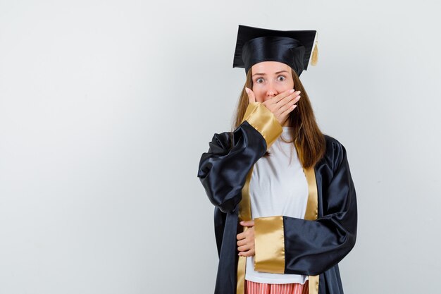 Porträt des weiblichen Absolventen, der Hand auf Mund im akademischen Kleid hält und schockierte Vorderansicht schaut