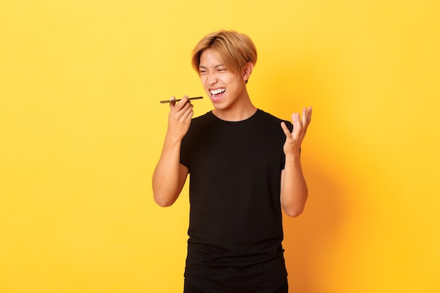 Porträt des wütenden asiatischen Mannes, der wütend auf Handy-Lautsprecher schreit, wütende Sprachnachricht aufzeichnen, gelbe Wand stehend