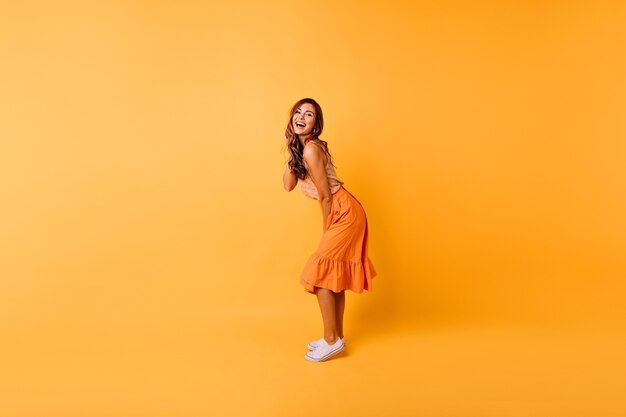 Porträt des wunderschönen lockigen Mädchens im orangefarbenen Rock in voller Länge. Studioporträt der positiven Dame, die auf Gelb tanzt.