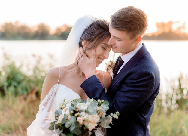 Porträt des zarten Hochzeitspaares nahe Wasser, das fast mit schönem Hochzeitsstrauß in den Händen am warmen Abend küsst