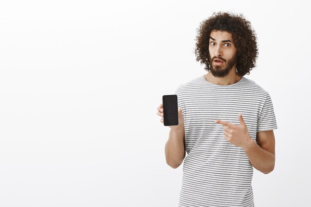 Porträt des zweifelhaften ungläubigen attraktiven Mannes mit lockigem Haar, hält Smartphone und zeigt mit Zeigefinger auf Gerät