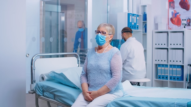 Porträt einer älteren Dame, die während der COVID-19-Krise auf dem Krankenhausbett sitzt. Arzt und Patienten mit Schutzmaske und Ausrüstung in moderner Privatklinik. Gesundheitssystem nach Coronavirus