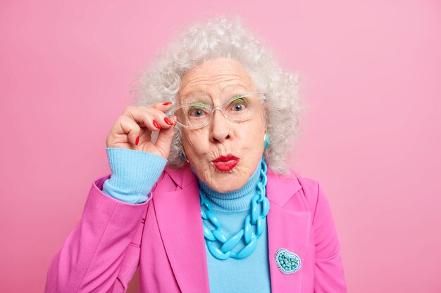 Porträt einer älteren europäischen Frau mit lockigem grauem Haar hält die Hand am Brillenrand und hält die Lippen in modischer Kleidung abgerundet