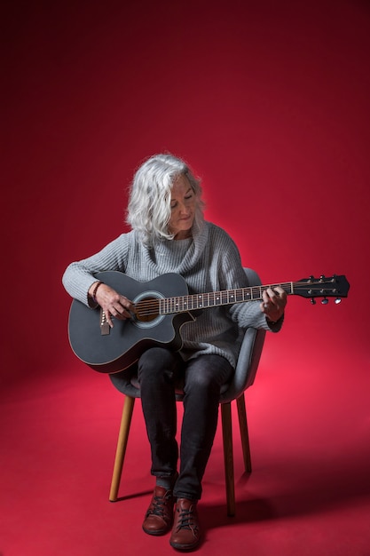 Porträt einer älteren Frau, die auf dem Stuhl spielt die Gitarre gegen roten Hintergrund sitzt