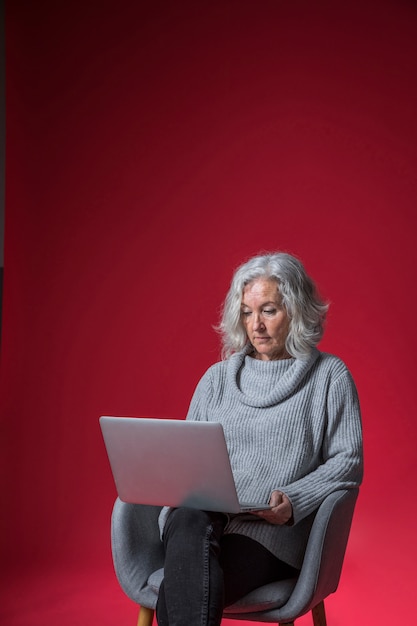 Porträt einer älteren Frau, die auf Lehnsessel unter Verwendung des Laptops gegen roten Hintergrund sitzt