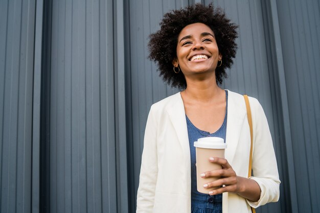 Porträt einer afro-geschäftsfrau, die eine tasse kaffee hält, während sie draußen auf der straße steht