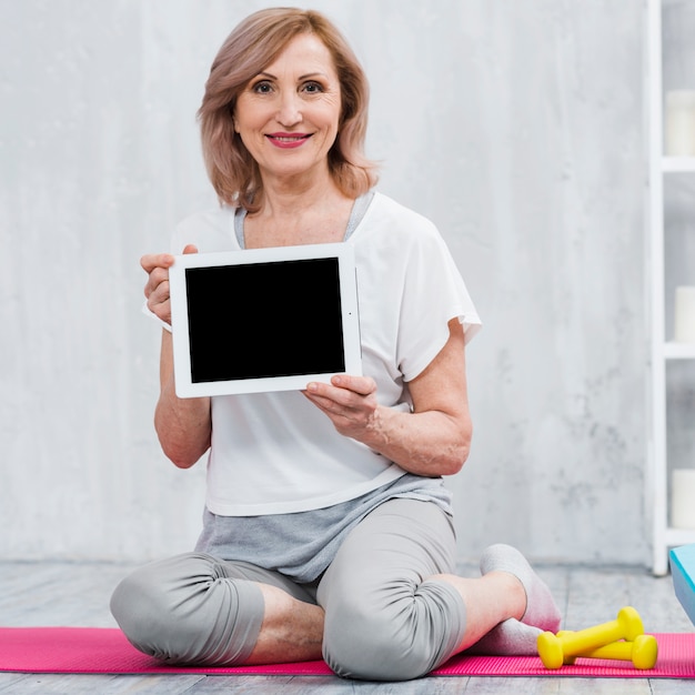 Porträt einer alten frau, die auf der yogamatte in der hand hält digitale tablette sitzt