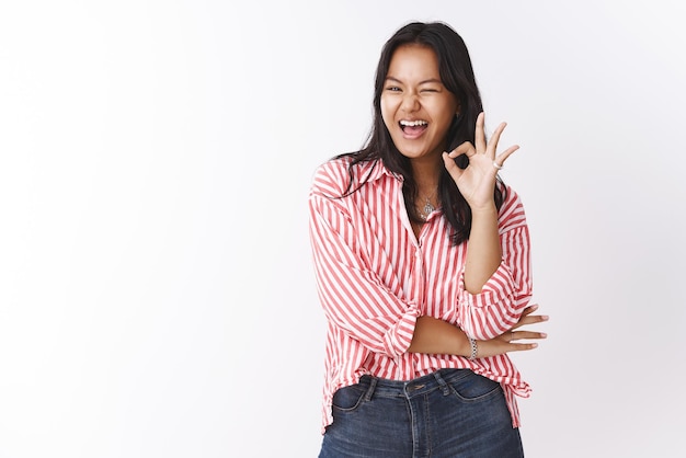Porträt einer amüsierten und beeindruckten optimistischen, stilvollen asiatischen Frau, die zwinkert und ein okayzeichen zeigt