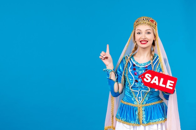 Porträt einer aserbaidschanischen Frau in traditioneller Kleidung, die das Typenschild des Verkaufs auf blauem Hintergrund hält, ethnische Tänzerin, die Frühlings-Shopping-Foto