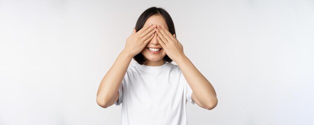 Porträt einer asiatischen Frau, die die Augen bedeckt und auf eine Überraschung mit verbundenen Augen wartet, die glücklich vor dem weißen Hintergrund steht