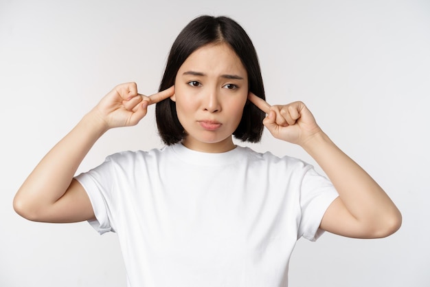 Porträt einer asiatischen Frau, die die Ohren schließt und sich durch laute Geräusche unwohl fühlt, die über weißem Hintergrund stehen