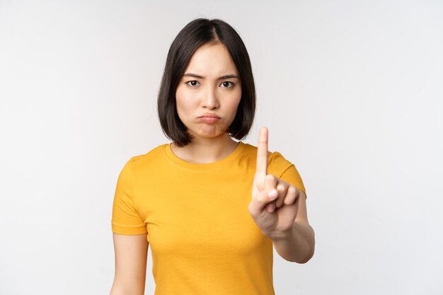 Porträt einer asiatischen Frau, die ernst und wütend aussieht und Stopp-Verbot-Gesten-Tabu-Schild zeigt, das smth verbietet, in gelbem T-Shirt auf weißem Hintergrund zu stehen