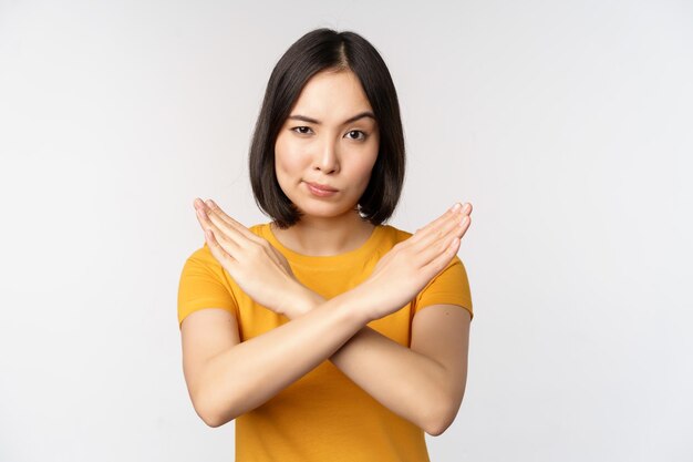 Porträt einer asiatischen Frau, die ernst und wütend aussieht und Stopp-Verbot-Gesten-Tabu-Schild zeigt, das smth verbietet, in gelbem T-Shirt auf weißem Hintergrund zu stehen