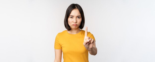 Porträt einer asiatischen Frau, die ernst und wütend aussieht und Stopp-Verbot-Gesten-Tabu-Schild zeigt, das smth verbietet, in gelbem T-Shirt auf weißem Hintergrund zu stehen