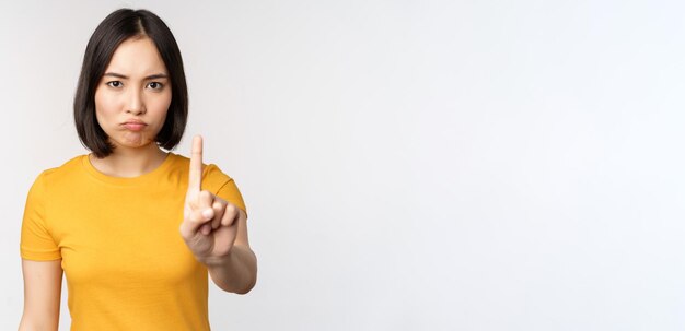 Porträt einer asiatischen Frau, die ernst und wütend aussieht und Stopp-Verbot-Gesten-Tabu-Schild zeigt, das smth verbietet, in gelbem T-Shirt auf weißem Hintergrund zu stehen