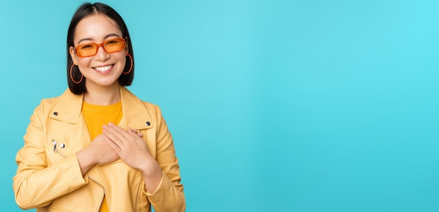 Porträt einer asiatischen Frau, die lächelnd Händchen auf dem Herzen hält und mit Zärtlichkeit in die Kamera schaut, dankbare Emotion, die über blauem Hintergrund steht