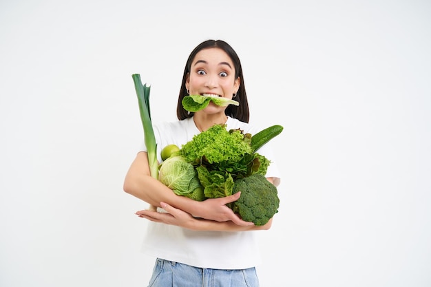 Porträt einer asiatischen Frau, die Salat isst und grünes Bio-Gemüse auf einem weißen Hintergrund hält