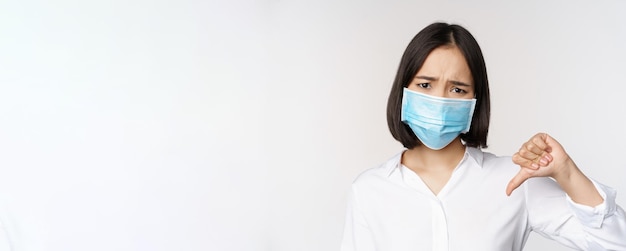 Porträt einer asiatischen Frau in medizinischer Gesichtsmaske, die Daumen nach unten zeigt, mit enttäuschten, müden Gesichtsausdrücken