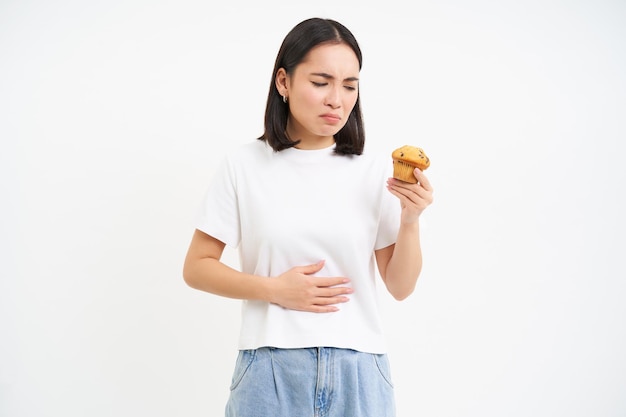 Porträt einer asiatischen Frau mit Bauchschmerzen, die Cupcake in der Hand hält, Lebensmittelvergiftung und sich unwohl fühlt, Studiohintergrund