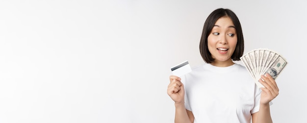 Porträt einer asiatischen Frau mit Gelddollar und Kreditkarte, die beeindruckt und verblüfft im Stehen ...