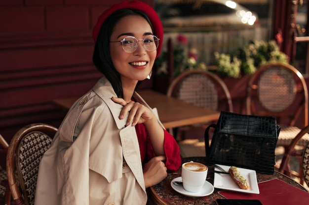 Porträt einer asiatischen gebräunten Frau in rotem Kleid, stilvollem Baskenmütze, beigem Trenchcoat und Brille sitzt in einem gemütlichen Straßencafé, lächelt und genießt Dessert und Kaffee