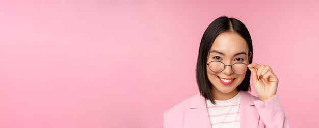 Porträt einer asiatischen Geschäftsfrau mit Brille, die fasziniert in die Kamera schaut und lächelnde professionelle Verkäuferin, die mit Interesse auf rosa Hintergrund starrt